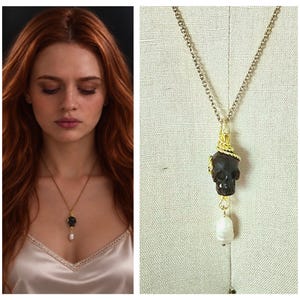Pode incluir: Um colar de corrente em tom dourado com um pingente de caveira escura esculpida envolto em fio dourado, com uma única pérola branca de formato irregular pendurada na base da caveira. O pingente está suspenso por uma delicada corrente.