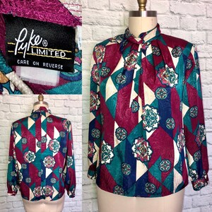 May include: A long-sleeved blouse with a geometric pattern in shades of burgundy, teal, white, and blue. The blouse has a high neck with a bow tie detail. The label reads "Lyke Limited, Care on Reverse."