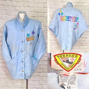 May include: A light blue denim jacket with a  "Jazzercise" logo on the back. The jacket has a stand-up collar, a button closure, and two large pockets on the front. The jacket is decorated with colorful stars and a gold patch. The jacket has a "Torpedo International" label on the bottom.