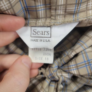 May include: Close-up of a vintage garment tag from Sears, Made in U.S.A. The tag is white with black text, and the garment is a tan plaid with blue and white accents. The tag also indicates the style number and size 18.