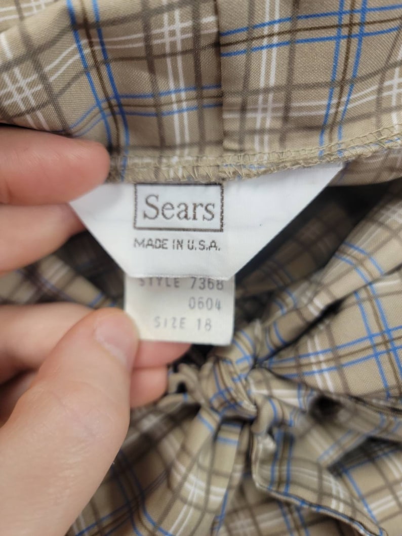 May include: Close-up of a vintage garment tag from Sears, Made in U.S.A. The tag is white with black text, and the garment is a tan plaid with blue and white accents. The tag also indicates the style number and size 18.