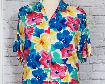 Könnte beinhalten: Ein kurzärmeliges Hemd mit Knöpfen und einem farbenfrohen Blumendruck. Das Hemd hat einen weißen Hintergrund mit großen rosa, blauen, gelben und grünen Blumen.