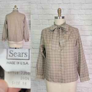 May include: A vintage Sears blouse in a plaid pattern, displayed on a mannequin. The long-sleeved shirt features a bow tie collar and button-down front. The tag reads "Sears MADE IN U.S.A." and "SIZE 18". The shirt is tan, blue, and white.