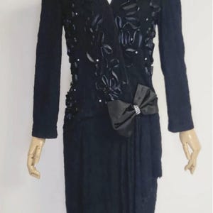 May include: A black dress with a fitted bodice and a skirt that falls to the knee. The bodice is adorned with black satin flowers and rhinestones. A large black satin bow with a rhinestone accent is tied at the waist.