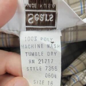 May include: Close-up of a clothing tag with the text "MADE IN USA" and the brand name "26912". The tag also includes washing instructions and size information. The fabric has a plaid pattern in tan, blue, and white.