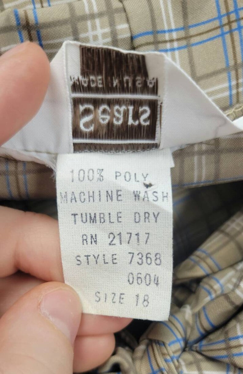 May include: Close-up of a clothing tag with the text "MADE IN USA" and the brand name "26912". The tag also includes washing instructions and size information. The fabric has a plaid pattern in tan, blue, and white.