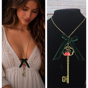 May include: A long gold-tone chain necklace featuring a vintage-style skeleton key pendant. The key is adorned with a dark green velvet ribbon bow and two small, coral-pink flower charms attached to the top of the key's bow section.