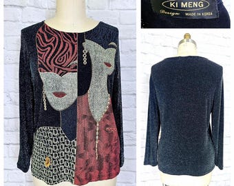 May include: A black long-sleeved shirt with a glittery texture. The front of the shirt features a colorful graphic design of two women's faces with gold accents. The back of the shirt is plain. The shirt has a label that reads "Ki Meng Design Made in Korea."