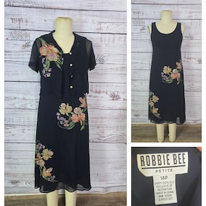Puede incluir: Un conjunto de vestido de dos piezas azul marino con bordados florales. El conjunto incluye un vestido sin mangas y una chaqueta abotonada de manga corta. El vestido y la chaqueta están adornados con flores bordadas de colores. La etiqueta dice "ROBBIE BEE PETITE 14P".