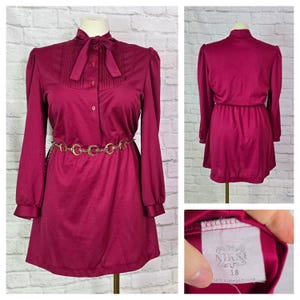 May include: A burgundy long-sleeved dress with a bow tie collar and a gold chain belt. The dress has a gathered waist and a label that reads "Sizes 18, Game & Company Ltd."