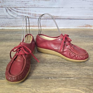 May include: A pair of vintage red leather shoes with matching red laces. The shoes have a rounded toe and a tan sole. The shoes are displayed on a wooden surface with a light-colored background.