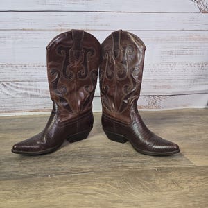 May include: A pair of brown leather cowboy boots with pointed toes and decorative stitching. The boots have a textured finish and a low heel, set against a wooden background.