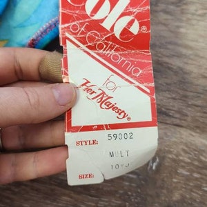 Pode incluir: Uma etiqueta vermelha e branca de "Cole of California for Her Majesty" com o n&uacute;mero de estilo 59002 e o tamanho 10VJ. A etiqueta &eacute; parcialmente vis&iacute;vel, com um tecido estampado colorido no fundo.