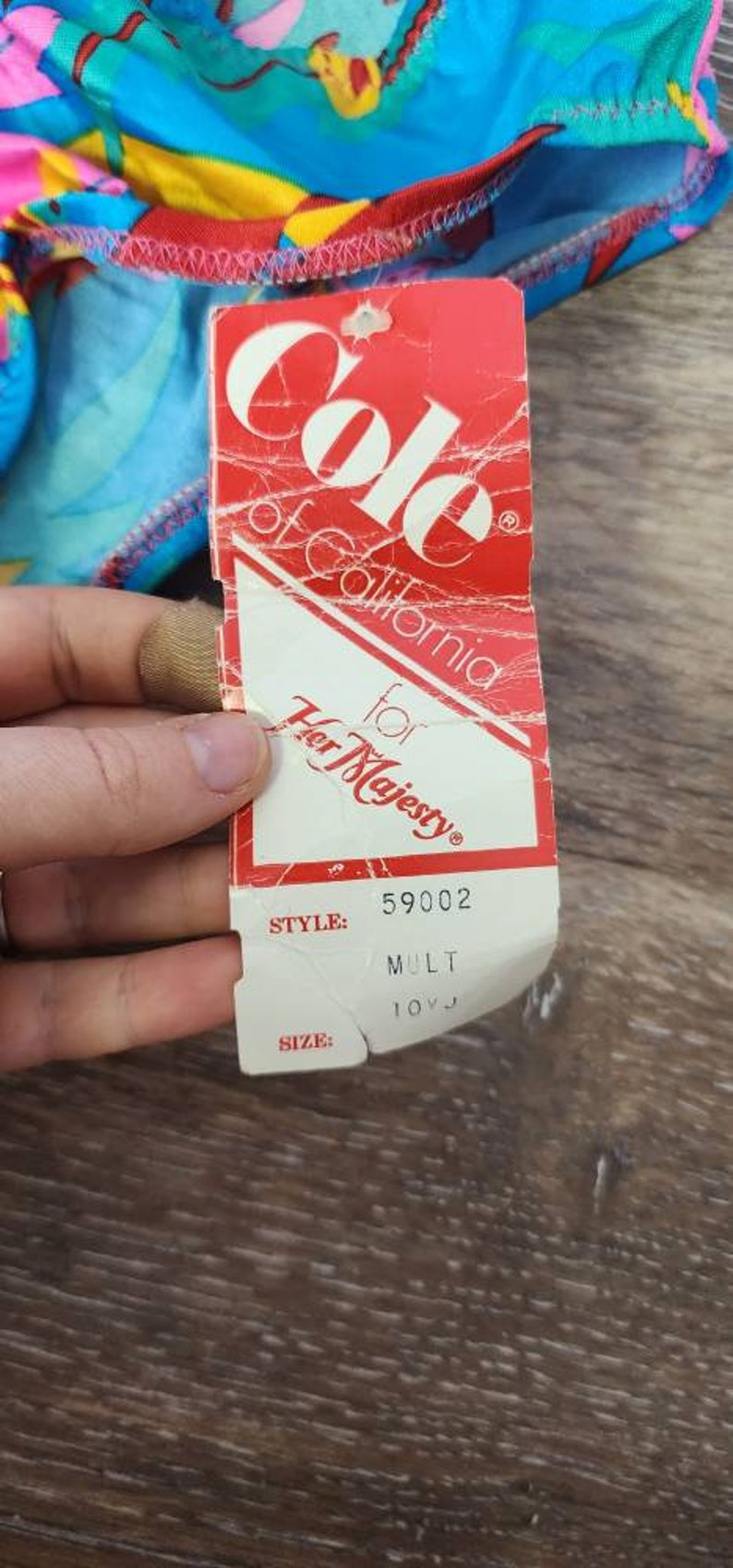 Pode incluir: Uma etiqueta vermelha e branca de "Cole of California for Her Majesty" com o n&uacute;mero de estilo 59002 e o tamanho 10VJ. A etiqueta &eacute; parcialmente vis&iacute;vel, com um tecido estampado colorido no fundo.