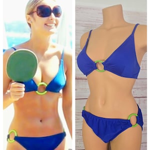 Może przedstawiać: Dwuczęściowy zestaw bikini w jednolitym królewskim niebieskim kolorze. Góra bikini ma trójkątny krój z cienkimi ramiączkami i jasnym, limonkowym kółkiem pośrodku. Pasujący dół od bikini posiada limonkową obręcz po bokach oraz kółka.