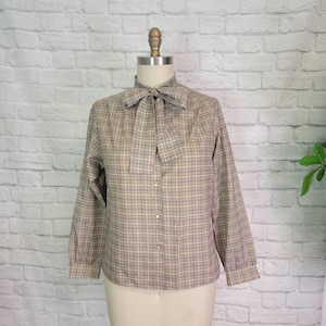 May include: A tan and blue plaid blouse with a necktie bow. The long-sleeved shirt has a button-down front and is displayed on a mannequin. The fabric has a subtle grid pattern, and the bow adds a classic touch to the garment.
