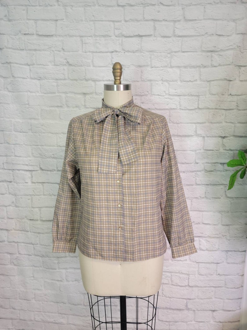 May include: A tan and blue plaid blouse with a necktie bow. The long-sleeved shirt has a button-down front and is displayed on a mannequin. The fabric has a subtle grid pattern, and the bow adds a classic touch to the garment.