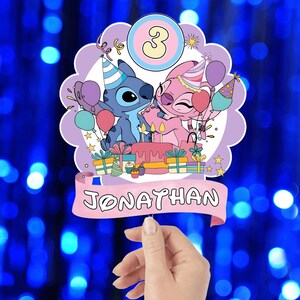 Może przedstawiać: Ozdoba na tort urodzinowy z postaciami Stitcha i Angel, z balonami, prezentami i tortem. Ozdoba ma cyfrę 3 i imię "Jonathan" na banerze. Tło jest rozmyte niebieskie.