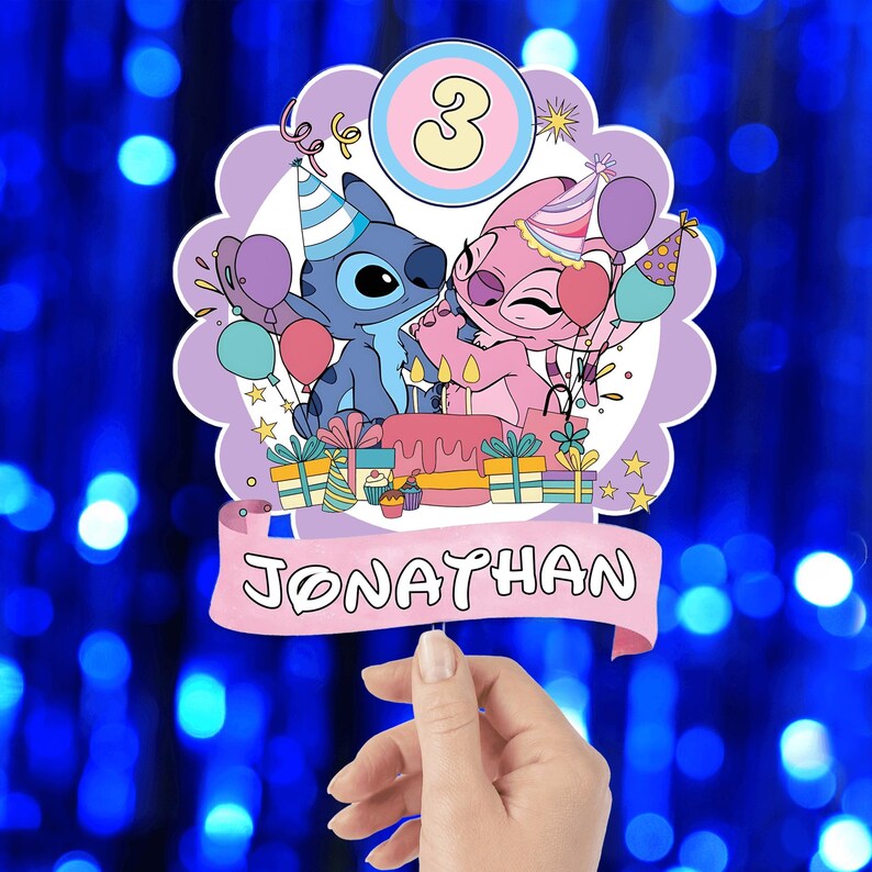 Może przedstawiać: Ozdoba na tort urodzinowy z postaciami Stitcha i Angel, z balonami, prezentami i tortem. Ozdoba ma cyfrę 3 i imię "Jonathan" na banerze. Tło jest rozmyte niebieskie.