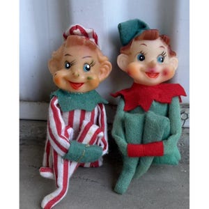 May include: Two vintage elf dolls with painted faces and rosy cheeks. The left elf wears a red and white striped outfit with a green collar and hat. The right elf wears a solid green outfit with a red collar and a matching green pointed hat.
