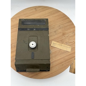 Puede incluir: Un dispositivo metálico verde oliva de forma rectangular, con un dial circular y una ventana de visualización rectangular. Una pequeña etiqueta de papel rectangular con texto está al lado del dispositivo. El dispositivo está sobre una superficie de madera.