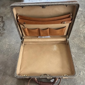 May include: An open briefcase with a textured brown and gray tweed exterior and a tan leather interior. The interior features a large main compartment, a wide document pocket, and three smaller utility pockets for organizing small items.