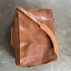 May include: A rectangular tan leather briefcase featuring a smooth front flap, a long adjustable shoulder strap with a reinforced pad, and a dark brown side handle. The leather surface displays natural grain textures and subtle surface markings.