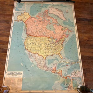 May include: A vintage map of North America, featuring detailed geographical information. The map is in shades of yellow, orange, and green, with text labels and a scale. The map is rolled up and hanging, with the title "North America" visible.