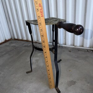 May include: A black metal trivet with a dark wooden handle, featuring a decorative openwork top and four legs. A wooden ruler is placed vertically against the trivet, showing a height measurement of approximately 17 inches from base to top.
