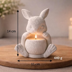 May include: A white ceramic bunny candle holder with a lit candle inside. The bunny is 14 cm tall and 11 cm wide, weighing 440 g. The candle holder is sitting on a wooden round tray.