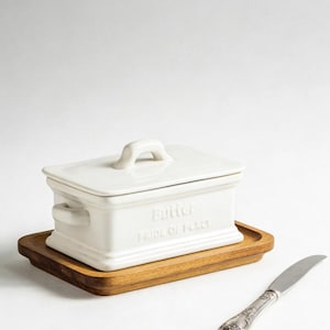 May include: A white ceramic butter dish with a lid and handle, inscribed with "Butter PRIDE OF PLACE". It sits on a wooden tray, with a silver butter knife to the side. The butter dish has a rectangular shape.