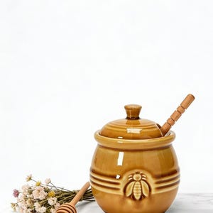 May include: A ceramic honey pot with a lid and a wooden honey dipper. The pot is brown with a raised bee design on the front. A small bouquet of dried flowers is next to the pot.