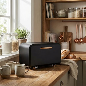May include: A black bread box with a wooden handle and feet sits on a wooden countertop. The kitchen scene includes jars of ingredients, mugs, and loaves of bread. A window and wooden shelves are in the background.
