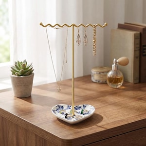 May include: A gold-colored jewelry stand with a wavy top bar and a heart-shaped base. The base is white with a blue floral pattern. Several necklaces, earrings, and a bracelet hang from the stand. A small succulent plant in a pot is visible in the background.