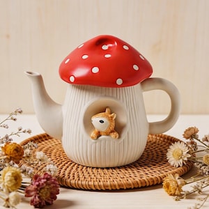 May include: A whimsical ceramic teapot shaped like a mushroom with a red cap and white polka dots. The teapot has a cream-colored body with a small squirrel figurine in a recessed area. It sits on a woven coaster, surrounded by dried flowers.