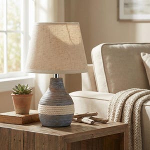 May include: A decorative table lamp with a beige linen shade and a ceramic base. The base is a mix of blue, gray, and brown tones, with a white rope accent. The lamp is on a wooden side table.
