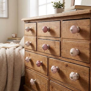 May include: A wooden dresser with multiple drawers, each adorned with a unique, decorative pink and white ceramic knob. The knobs vary in design, adding a touch of elegance to the rustic, aged wood.