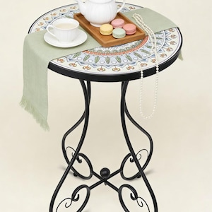 May include: A round, decorative side table with a mosaic tile top featuring a floral design in shades of blue, orange, and green. The table has a black wrought iron base. A tea set, macarons, and a pearl necklace are on the table.