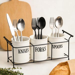 May include: A white ceramic utensil holder with three compartments labeled "KNIVES", "FORKS", and "SPOONS". The holder contains silverware and sits in a black metal frame. A wooden cutting board and a loaf of bread are in the background.