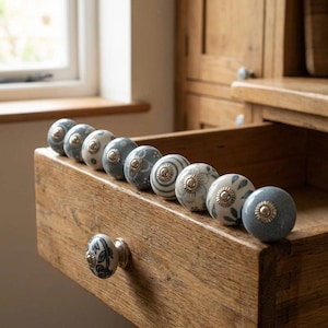 May include: A collection of decorative ceramic drawer pulls in various shades of blue and white, with floral patterns and metallic accents. The pulls are arranged in a wooden drawer, showcasing their design and texture.