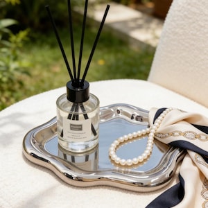 May include: A clear glass diffuser bottle with black reeds sits on a silver tray, next to a pearl necklace and a cream-colored scarf with navy and gold accents. The tray has a wavy edge.