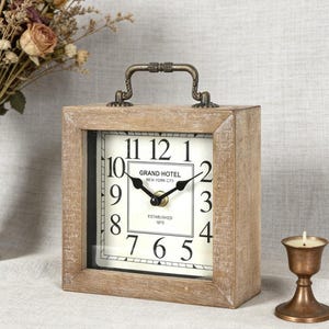 May include: A rectangular wooden clock with a square face. The clock face is white with black numerals and hands. The words "GRAND HOTEL NEW YORK CITY ESTABLISHED 1870" are printed on the face. A metal handle is on top.