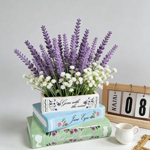 May include: A decorative floral arrangement featuring lavender and white flowers in a rectangular planter atop three ceramic book-shaped containers. The books are titled "Gone with the Wind", "Jane Eyre", and "Pride and Prejudice". A small white pitcher and a wooden calendar are also present.