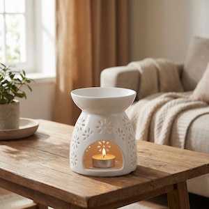 May include: A white ceramic essential oil diffuser with a floral cut-out design. A lit tea light candle is inside the diffuser, casting a warm glow. The diffuser sits on a wooden table, with a plant and a sofa in the background.