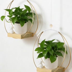 May include: Two hanging plant shelves. Each shelf features a gold-colored metal frame in a circular shape, a light wood shelf, and a white ceramic pot with a green plant. A small white figurine sits on each shelf.