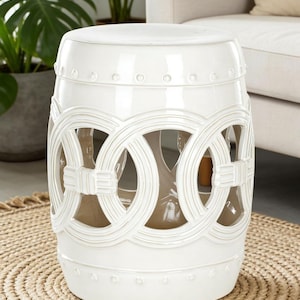May include: A white ceramic garden stool with a drum shape. The stool features a decorative openwork design of interlocking circles around the sides and a smooth, rounded top. The stool is placed on a round, woven mat.