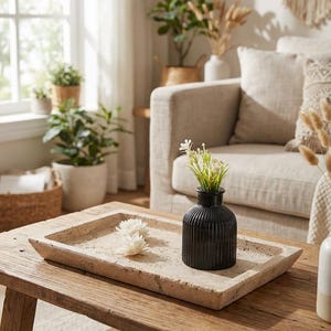 May include: A rustic wooden coffee table holds a rectangular stone tray with a black ribbed vase containing greenery and two white decorative flowers. The room features a neutral color palette with plants and a beige sofa.