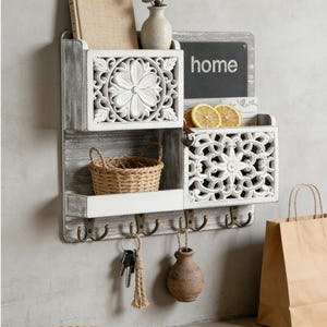 May include: A rustic, wall-mounted organizer with a distressed gray finish. It features carved white floral designs, a small chalkboard with the word "home", and several hooks. Includes a small woven basket and a paper bag.