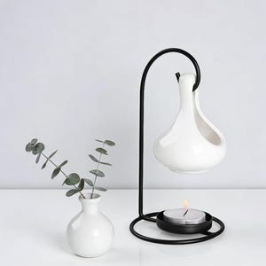 May include: A white ceramic essential oil diffuser with a black metal stand, holding a lit tea light candle. A small white vase with eucalyptus sprigs is to the left. The diffuser has a teardrop shape.