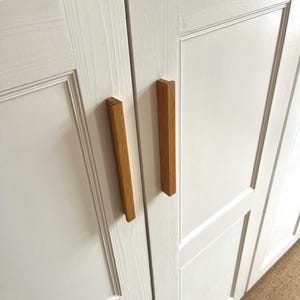May include: Close-up of a white cabinet with two wooden rectangular handles. The cabinet doors have a recessed panel design. The handles are a light brown colour and are vertically oriented.